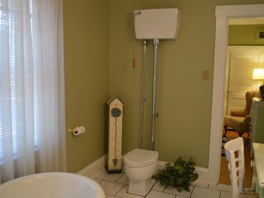 Bathroom with a tall toilet, white tub, sheer curtains, and doorway into another room. Olive-green walls and white trim.