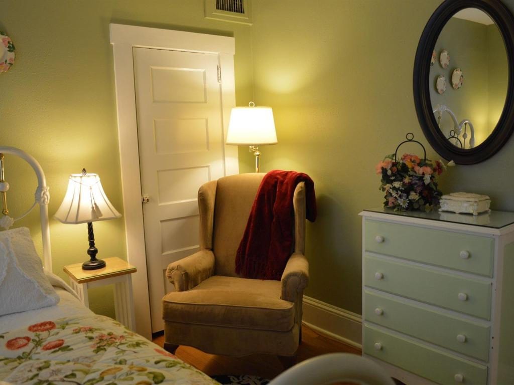 Cozy bedroom with a tan armchair, burgundy throw, dresser, round mirror, and a bed, painted in shades of green and cream.
