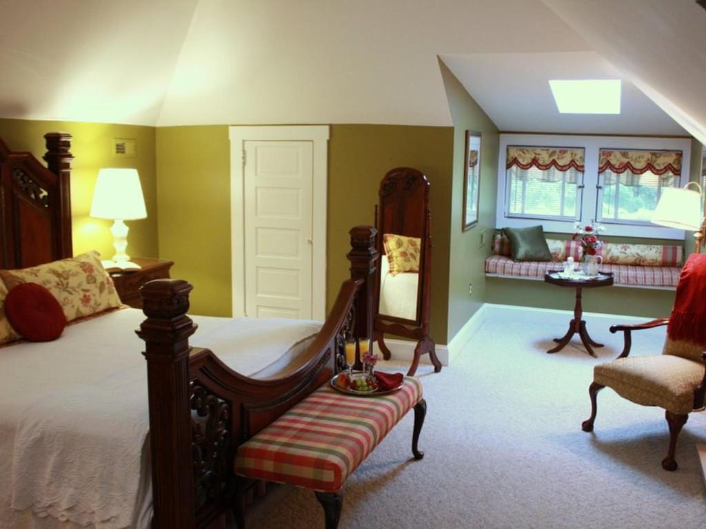 Bedroom with ornate wooden bed, bench, and tall mirror. The room has olive green walls, a window seat, and a skylight.