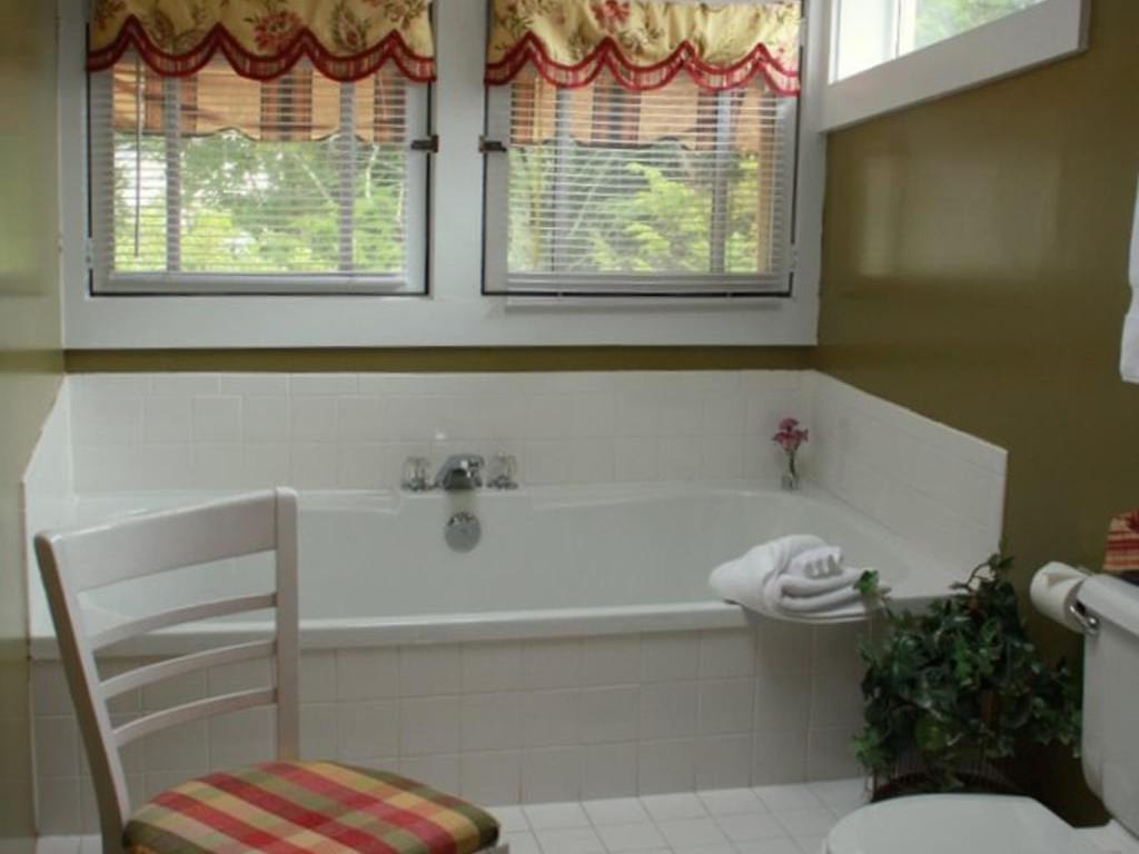 Bathroom with white-tiled bathtub, two windows with decorative curtains, a white chair, and a potted plant.