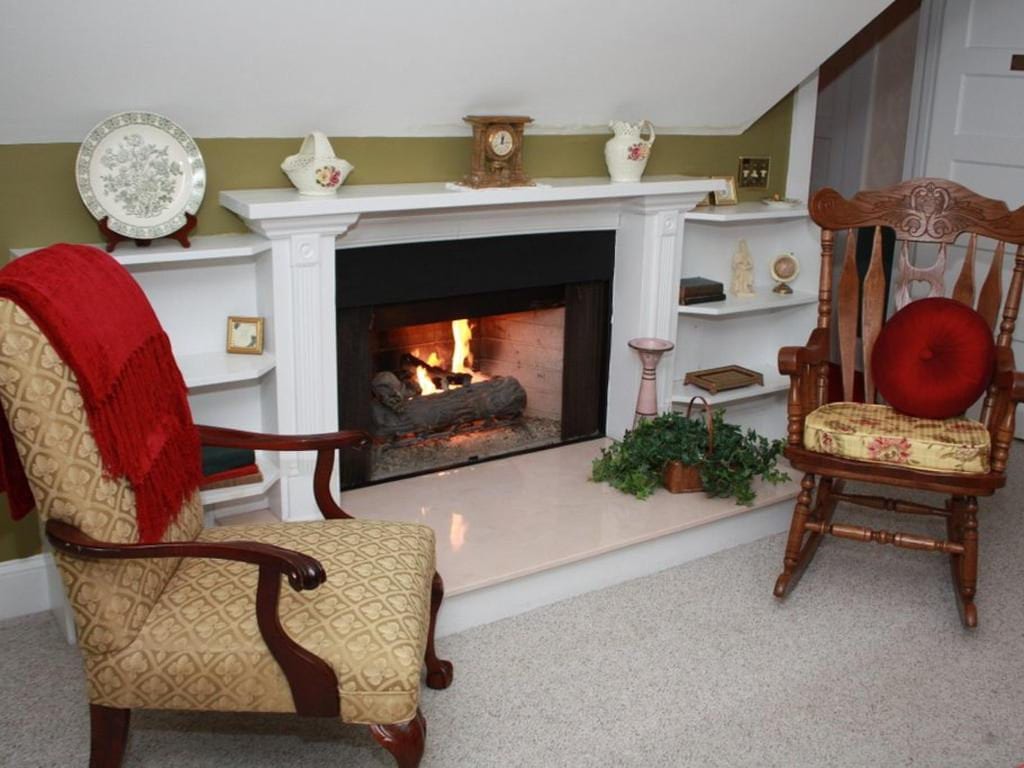 Cozy room with fireplace, armchair, and rocking chair. Red blanket draped over the armchair. Fire burning in the hearth.