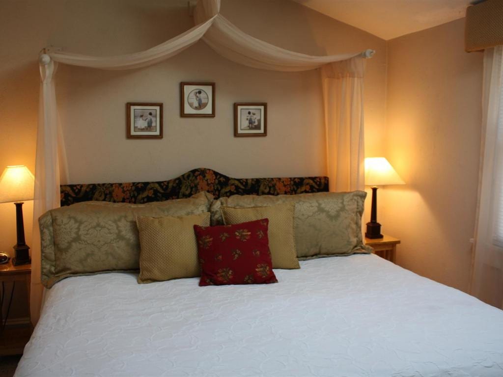 A bedroom with a bed featuring decorative pillows, two lamps, and framed pictures. The walls are painted tan.