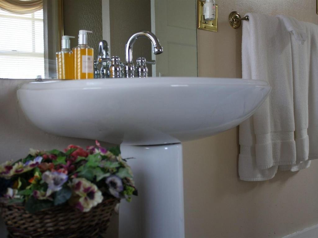 White pedestal sink with chrome faucet and soap dispensers. A basket of flowers sits in front. Towels hang on a nearby rack.