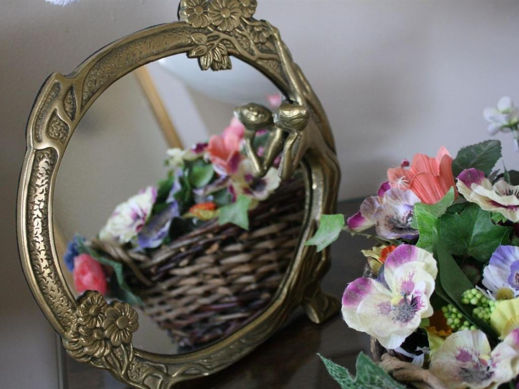 Oval brass-framed mirror reflecting a basket of flowers, beside a bouquet of colorful flowers.