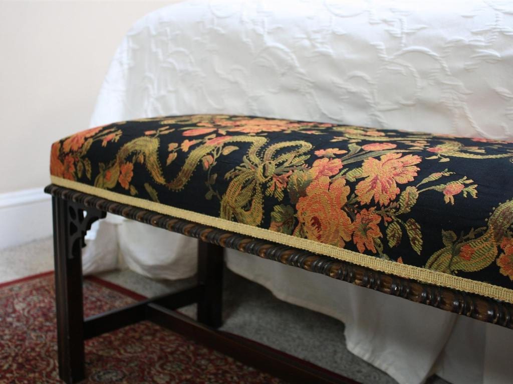 A dark wooden bench with a floral-patterned top, placed at the foot of a bed with a white comforter.