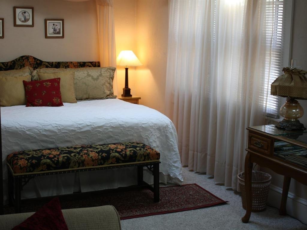 Cozy bedroom with a made bed, floral bench, sheer curtains, and a desk with a lamp next to a window.