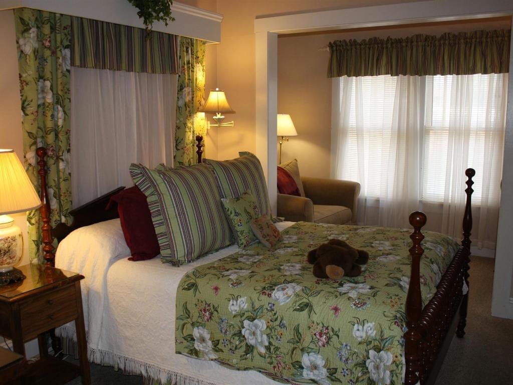 A bedroom with a floral bedspread, two lamps, and a view into another room with a chair and window.