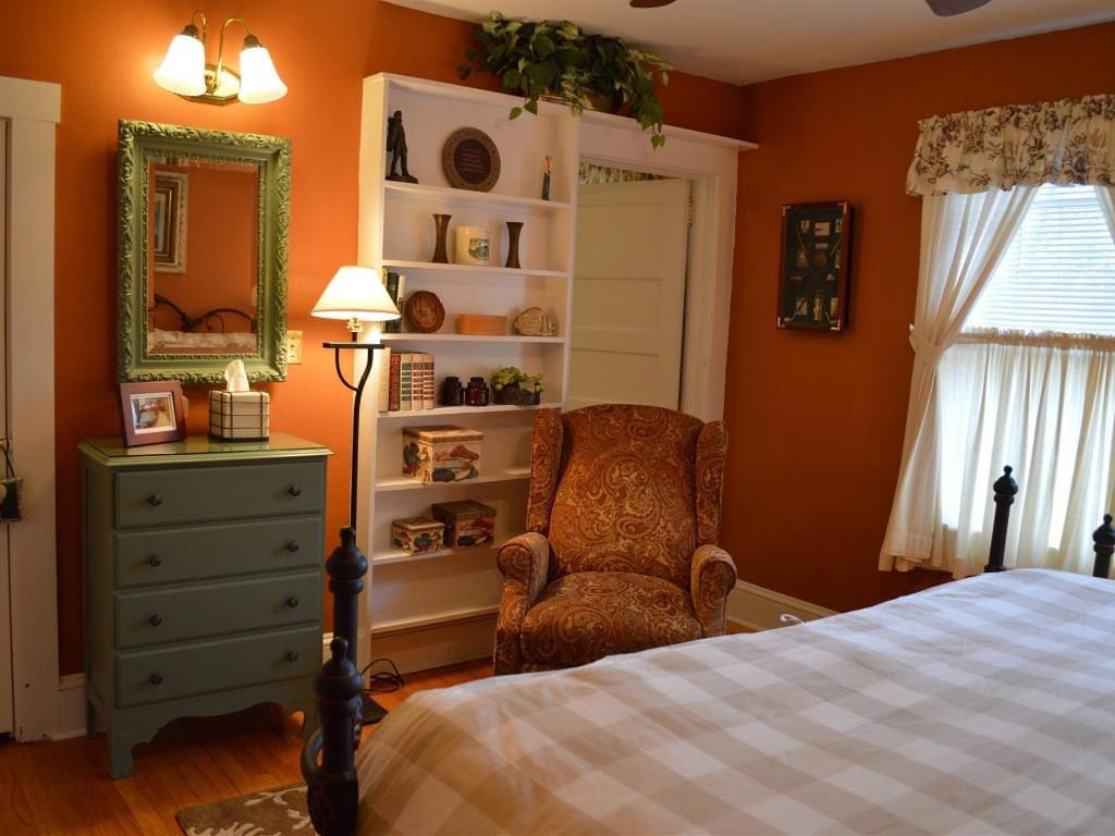 Cozy bedroom with orange walls, a bookshelf, a patterned armchair, and a bed with a floral-patterned curtain and a chest of drawers.