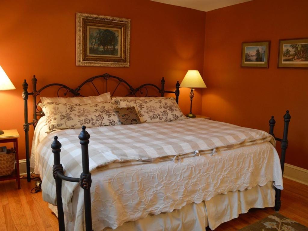 Bedroom with orange walls, iron bed frame, checkered bedspread, and framed artwork. Two lamps illuminate the bed.