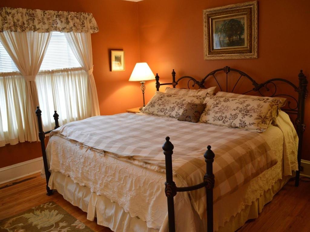 Cozy bedroom with a wrought iron bed, plaid bedding, and floral pillows. The room has orange walls, a window with cream curtains, and a lamp.