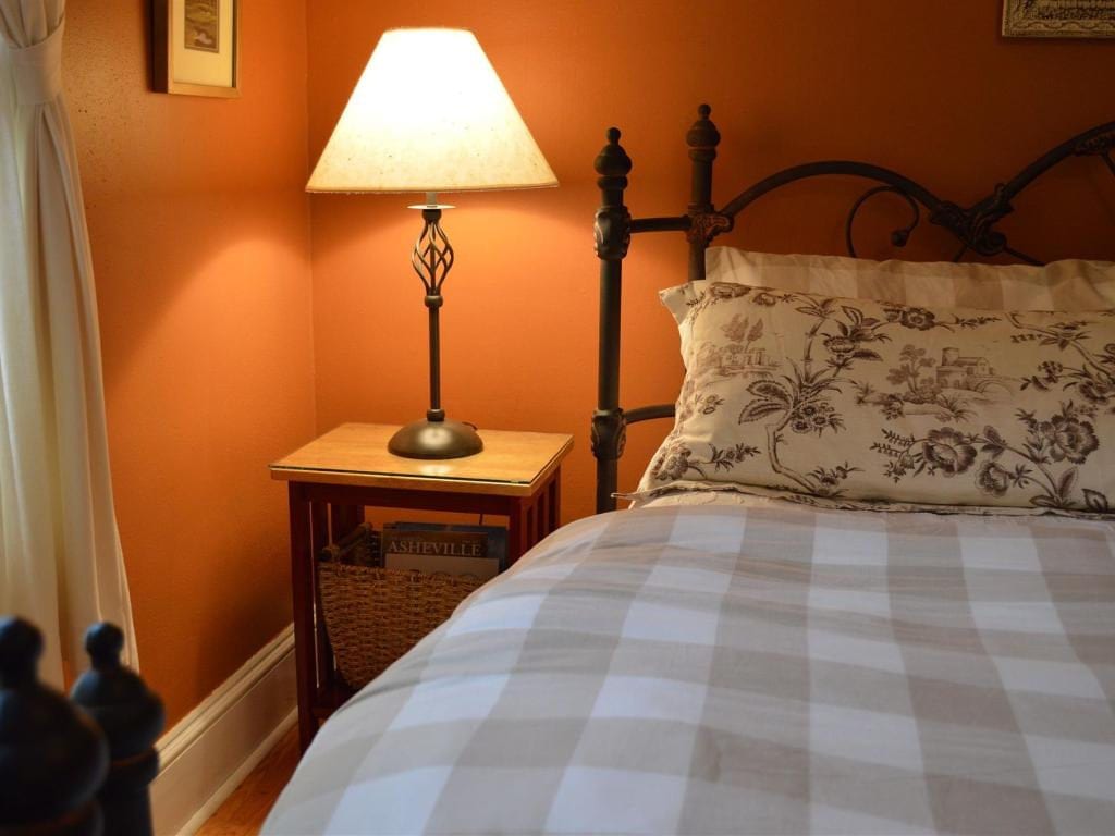 A lamp on a small wooden table next to a bed with a floral pillow and plaid comforter in a bedroom with orange walls.