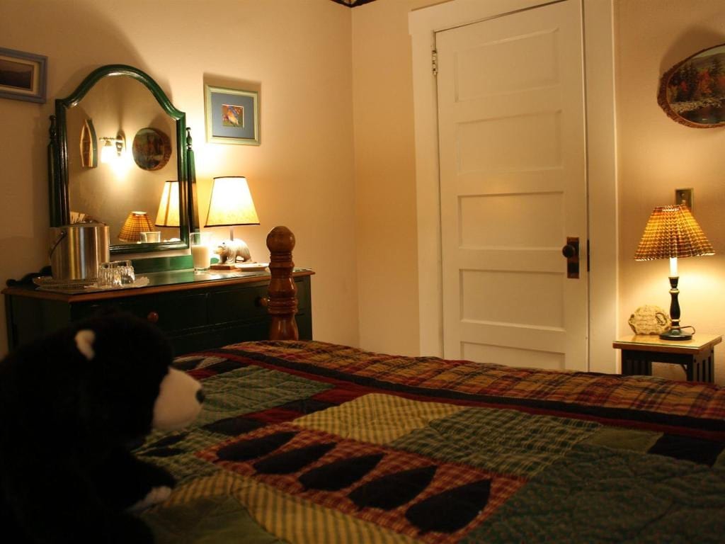Bedroom with a dark green dresser, bed with patchwork quilt, and a plush bear on the bed. Warm lighting creates a cozy atmosphere.