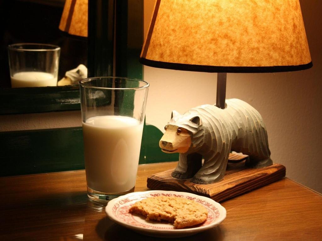 Glass of milk and half-eaten cookie on a plate next to a bear-shaped lamp on a nightstand.