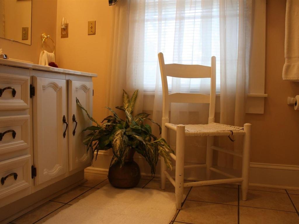 White wooden chair in front of a window with sheer curtains in a bathroom. A plant sits nearby next to a vanity.