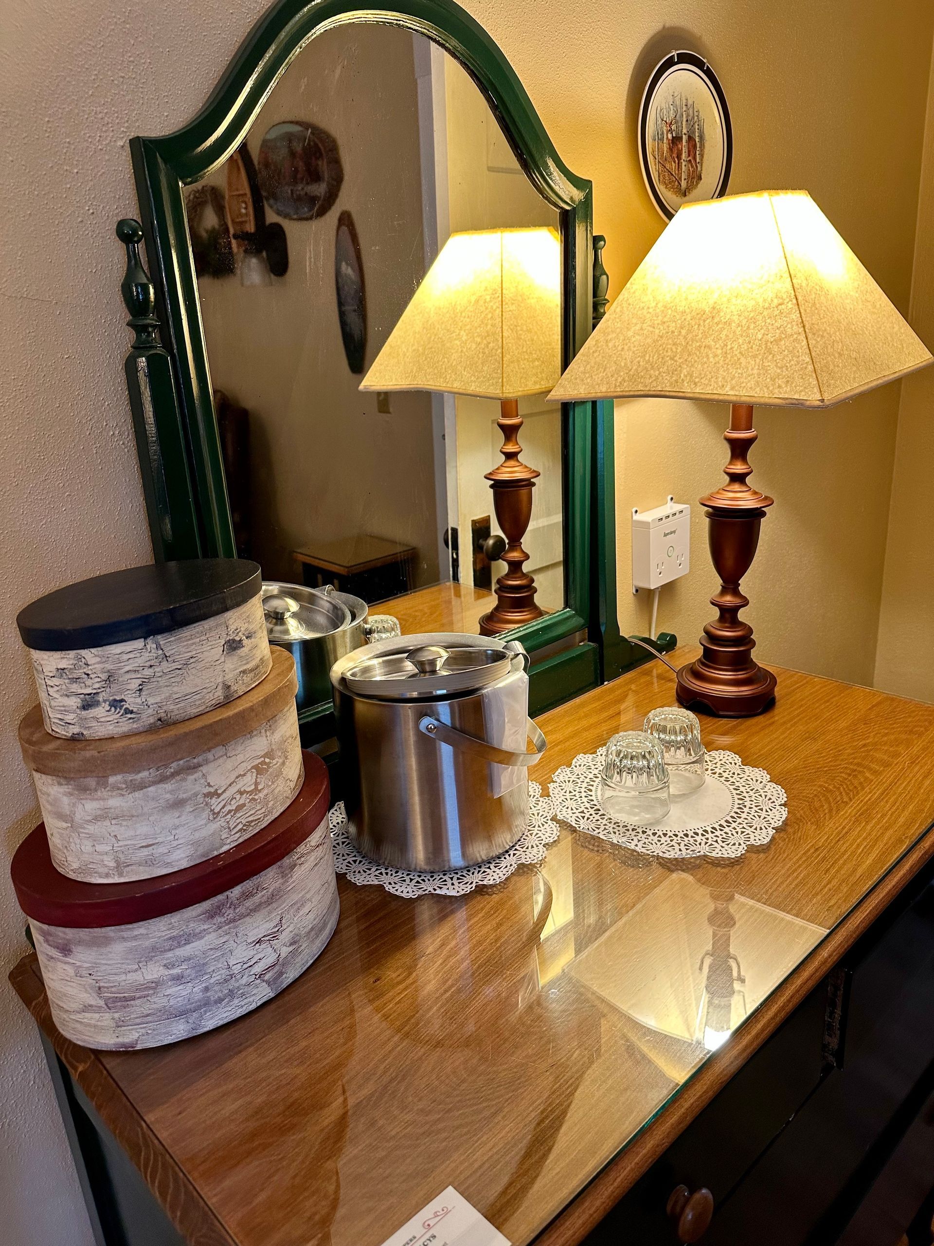 A dresser with a green-framed mirror, two lamps, decorative boxes, and a metal ice bucket, with reflections and warm lighting.