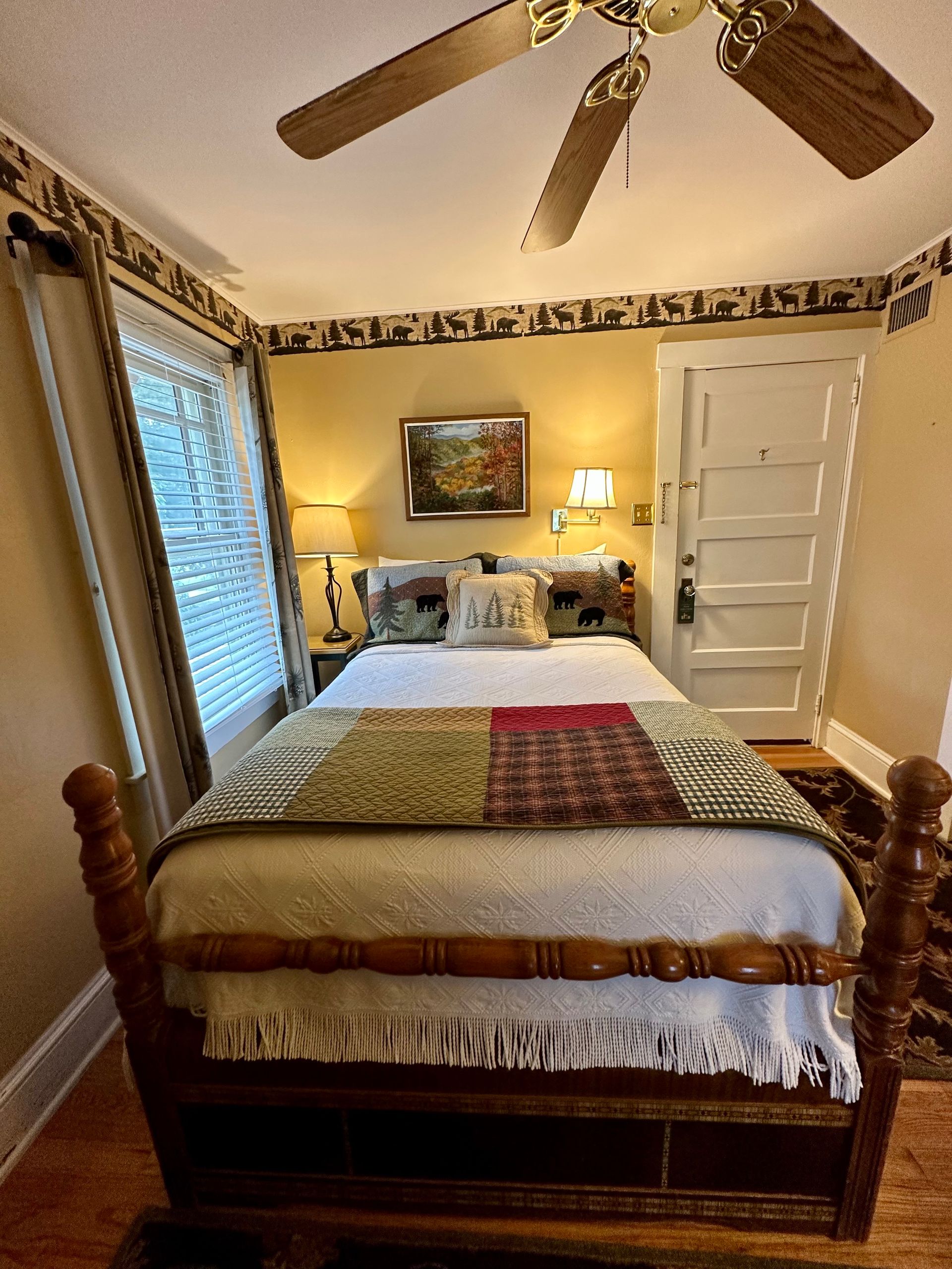 Bedroom with a four-poster bed, floral quilt, bedside lamps, and a window with blinds. Yellow walls and a wooden ceiling fan.