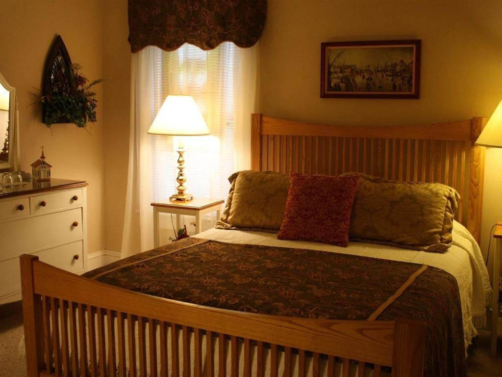 Cozy bedroom with a wooden bed frame, lamps, and a window with sheer curtains. Warm tones dominate.