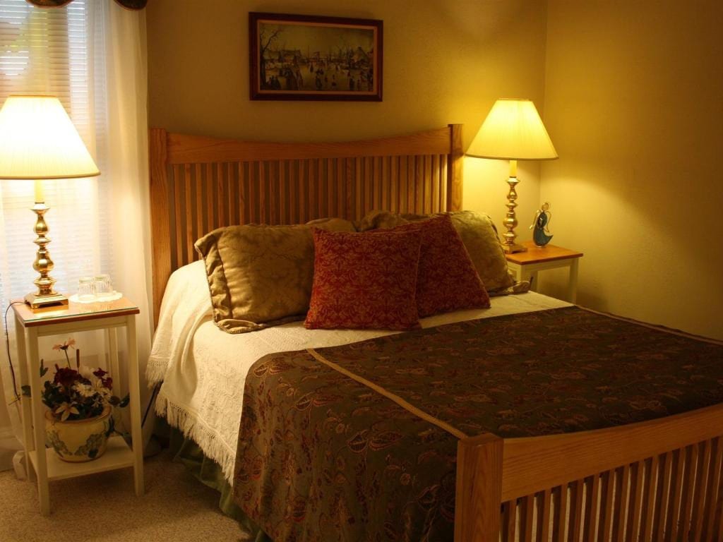 A cozy bedroom with a wooden bed frame, lamps on bedside tables, and decorative pillows. Warm tones and soft lighting.