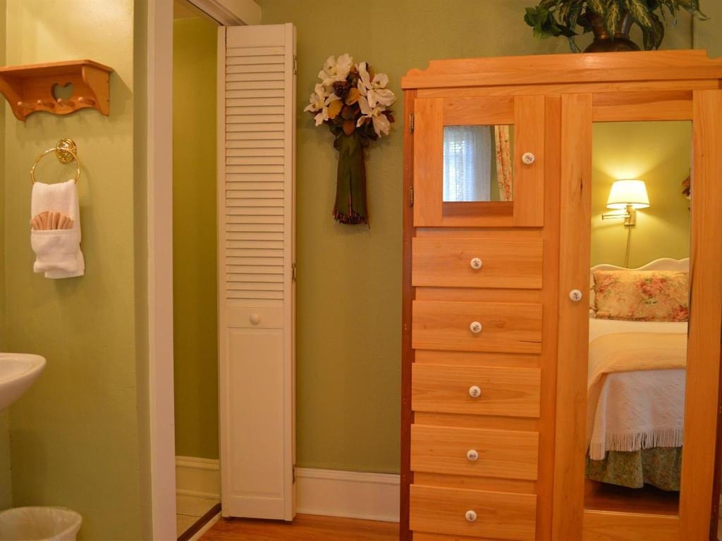 A bedroom with a wooden wardrobe and an open door to a bathroom with a sink. Green walls and a vase of flowers are present.