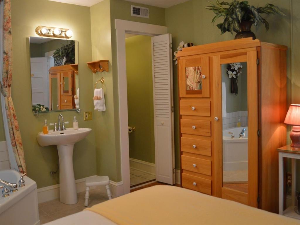 A bedroom with an attached bathroom. Light green walls, a wooden wardrobe, a pedestal sink, and a partially visible tub.