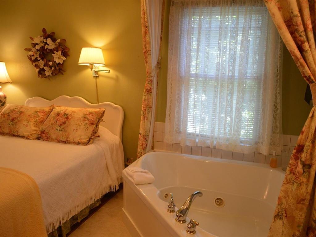 Cozy bedroom with a bed and a jacuzzi tub next to a window with lace curtains. The walls are painted a soft green.