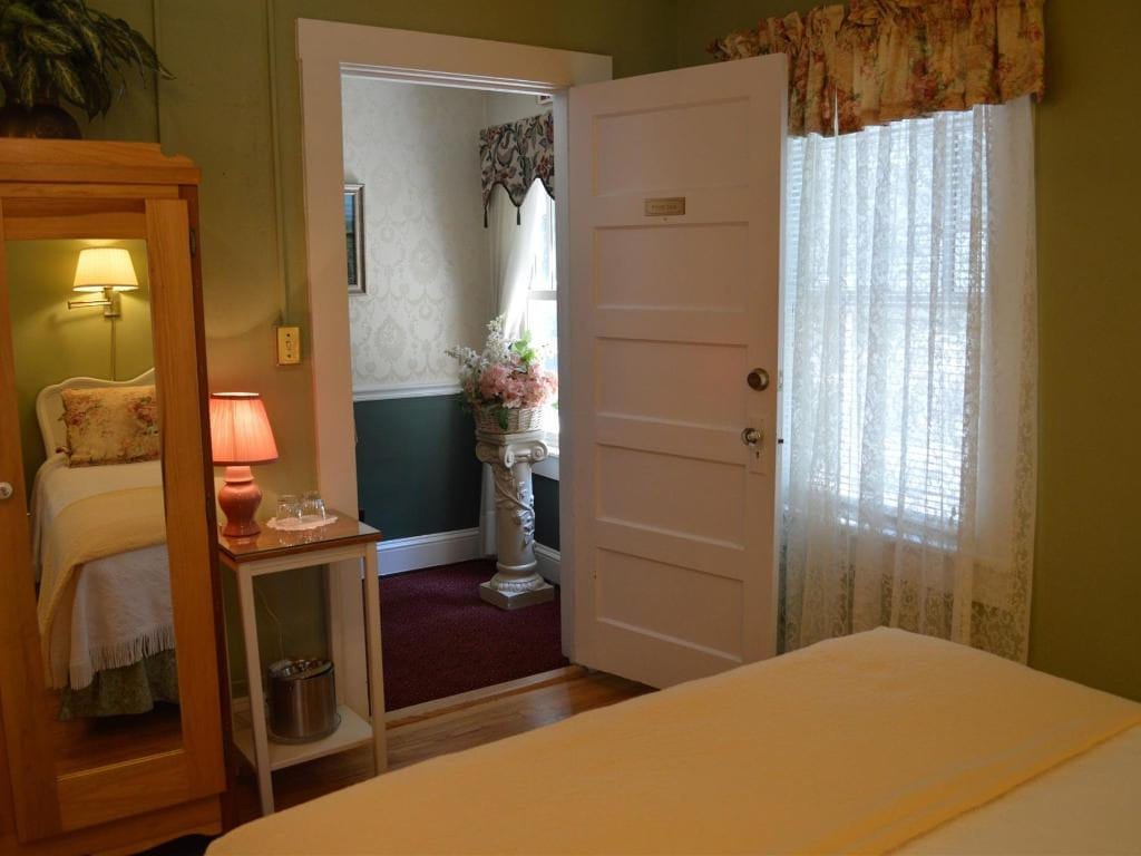 A bedroom interior with a bed, open door, and a mirror reflecting a bed. Pale yellow bedspread, green walls, and a glimpse of an interior room.