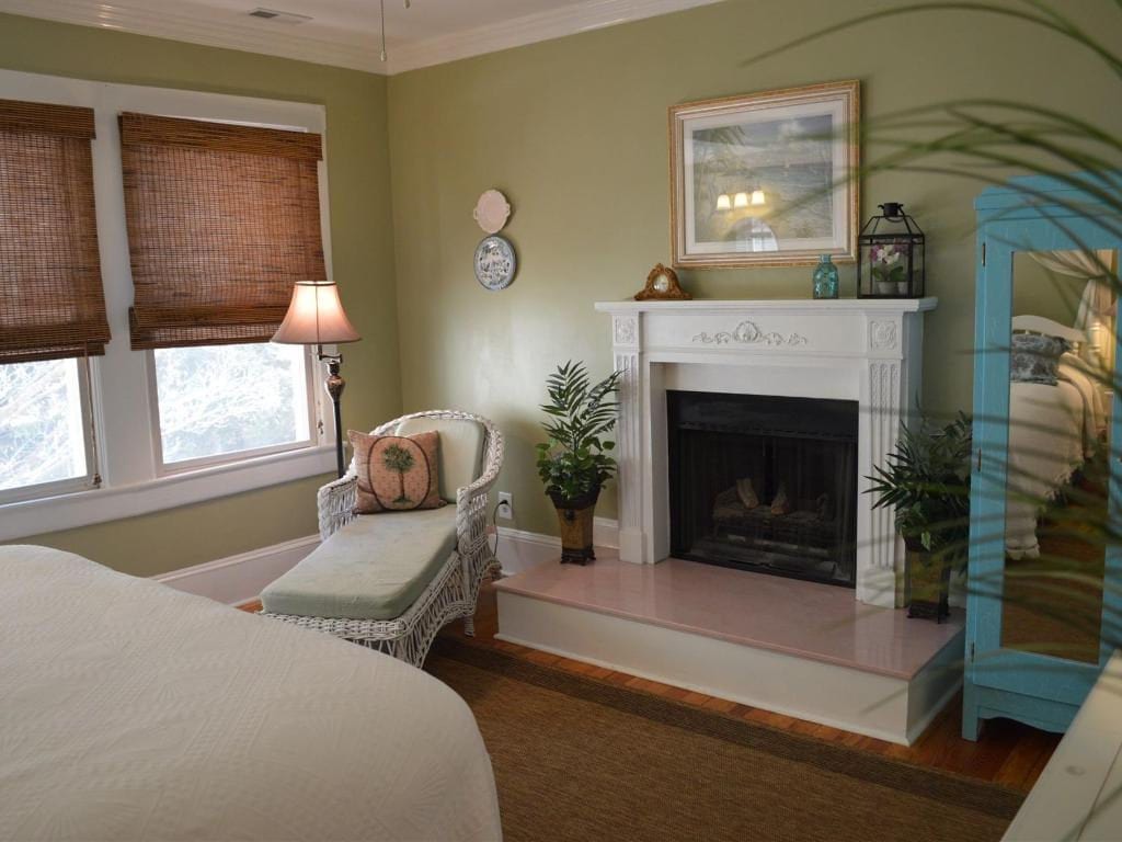 A bedroom with sage green walls, a white fireplace, a chaise lounge, and a mirror. Brown shades cover the windows.
