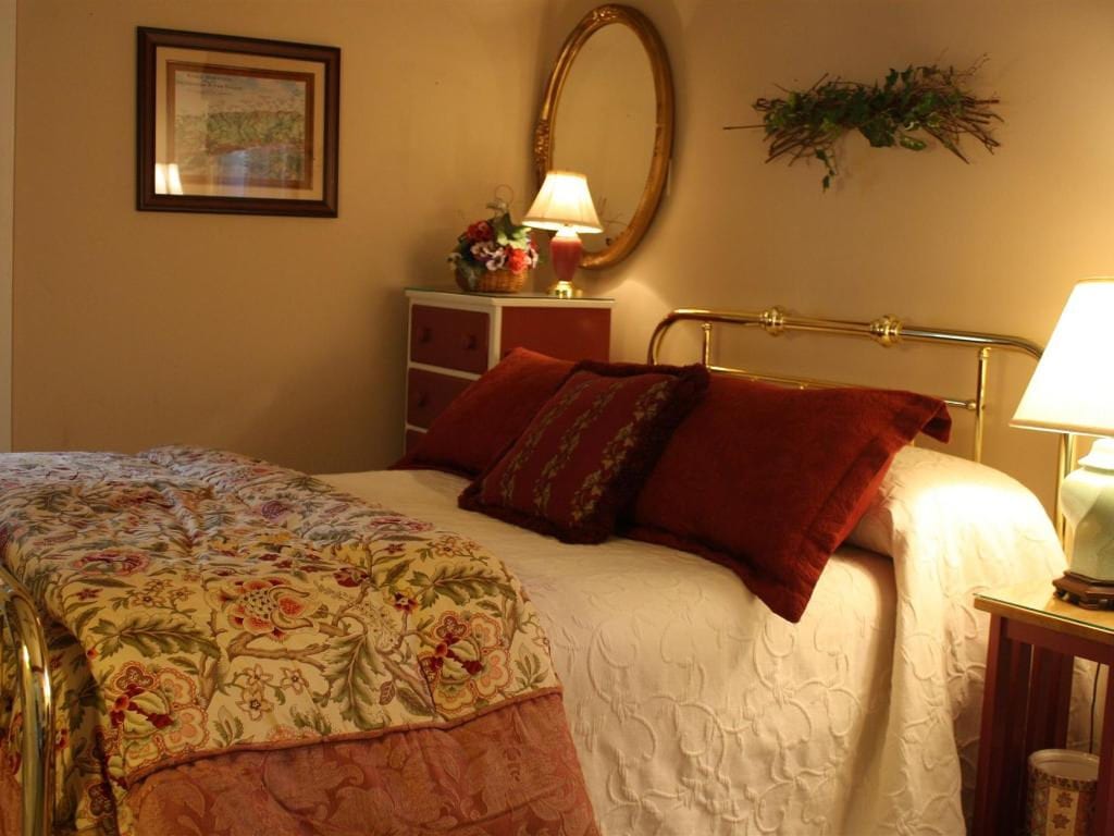 Bedroom with bed featuring floral bedding, red pillows, and a brass headboard. A dresser, mirror, and lamps are also visible.