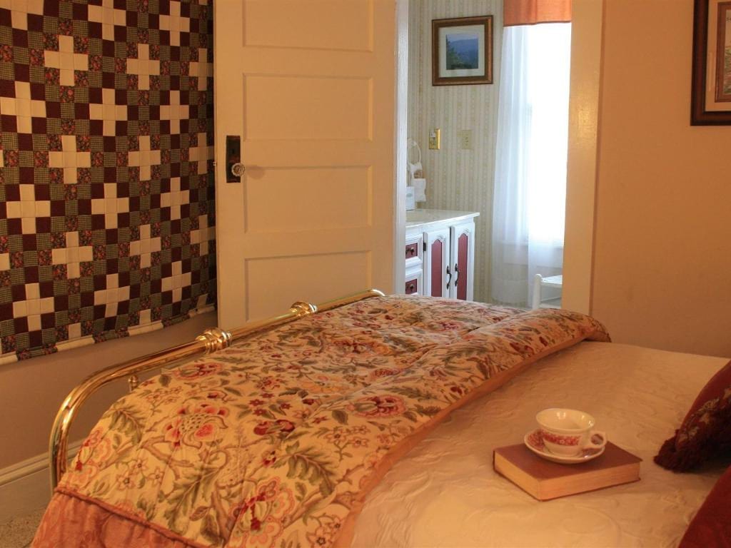 Bedroom with floral bedspread, brass bed frame, and a hanging quilt. An open doorway reveals a small bathroom.