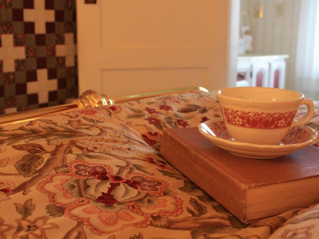 A teacup and saucer rest on a closed book atop a bed with floral sheets, near a quilt and open doorway.
