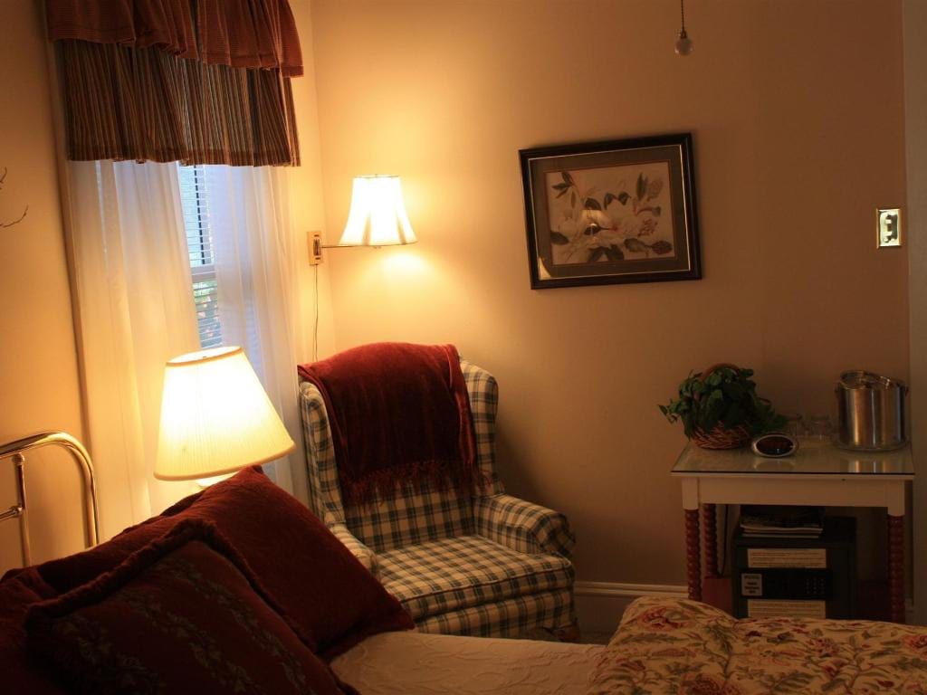 Cozy bedroom scene with a bed, plaid armchair, and small table. Warm lighting and a framed picture add to the inviting atmosphere.