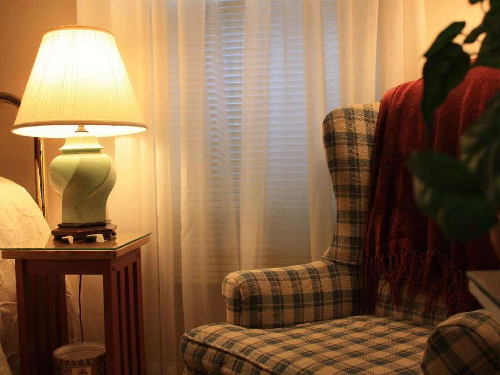 A lit lamp on a wooden table next to a plaid armchair with a red blanket; a window in the background.