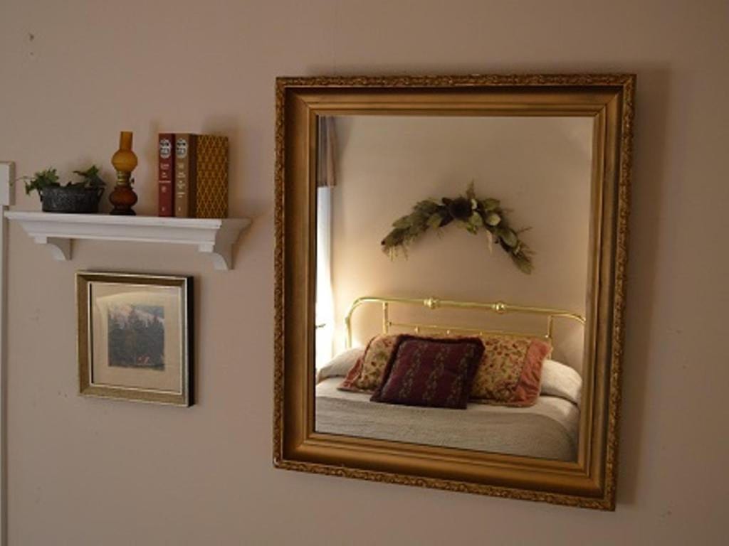 Mirror reflecting a bedroom with a gold headboard and decorative pillows, plus a shelf with books and a picture frame.
