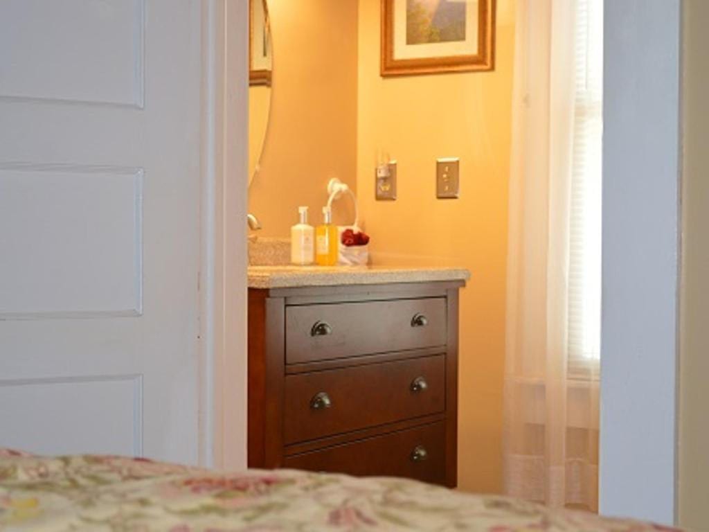 View through a doorway of a small bathroom with a dark wooden dresser under a sink, lit by sunlight.