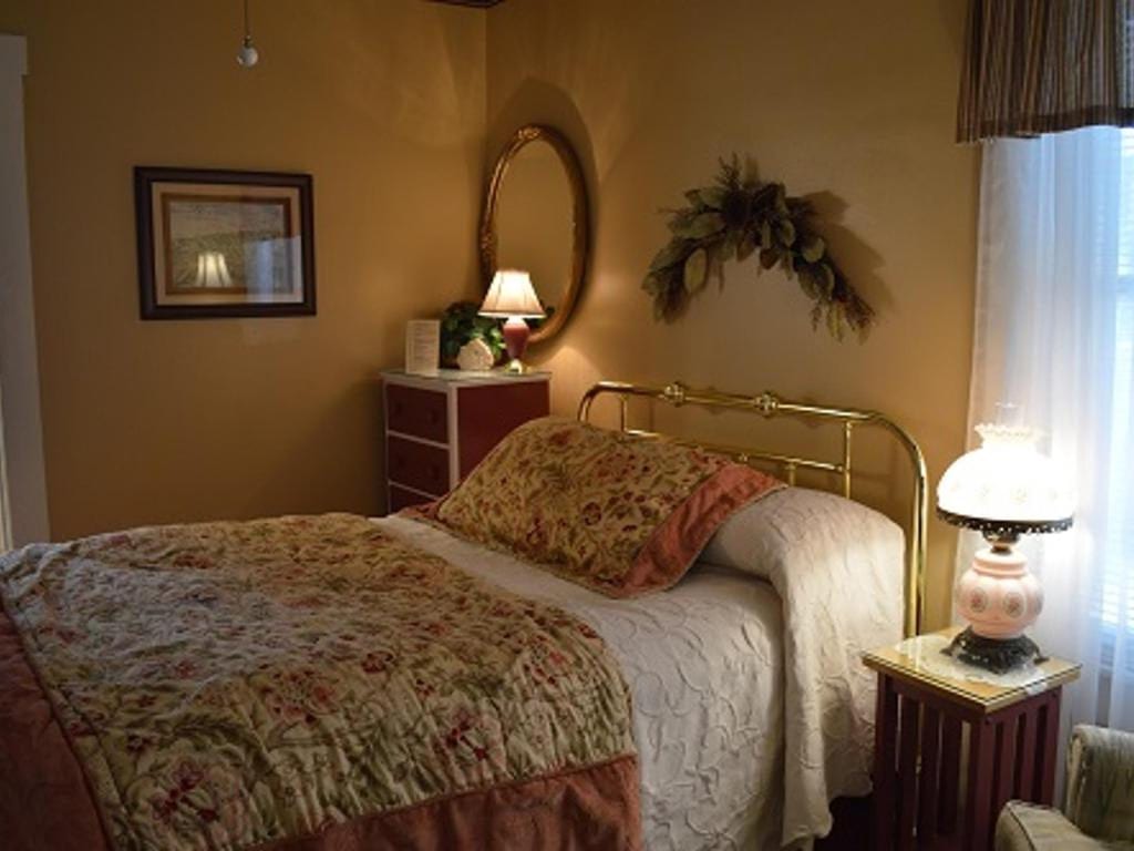 Cozy bedroom with a gold-colored bed frame, floral bedding, and a small nightstand with a lamp.  Warm colors and natural light.
