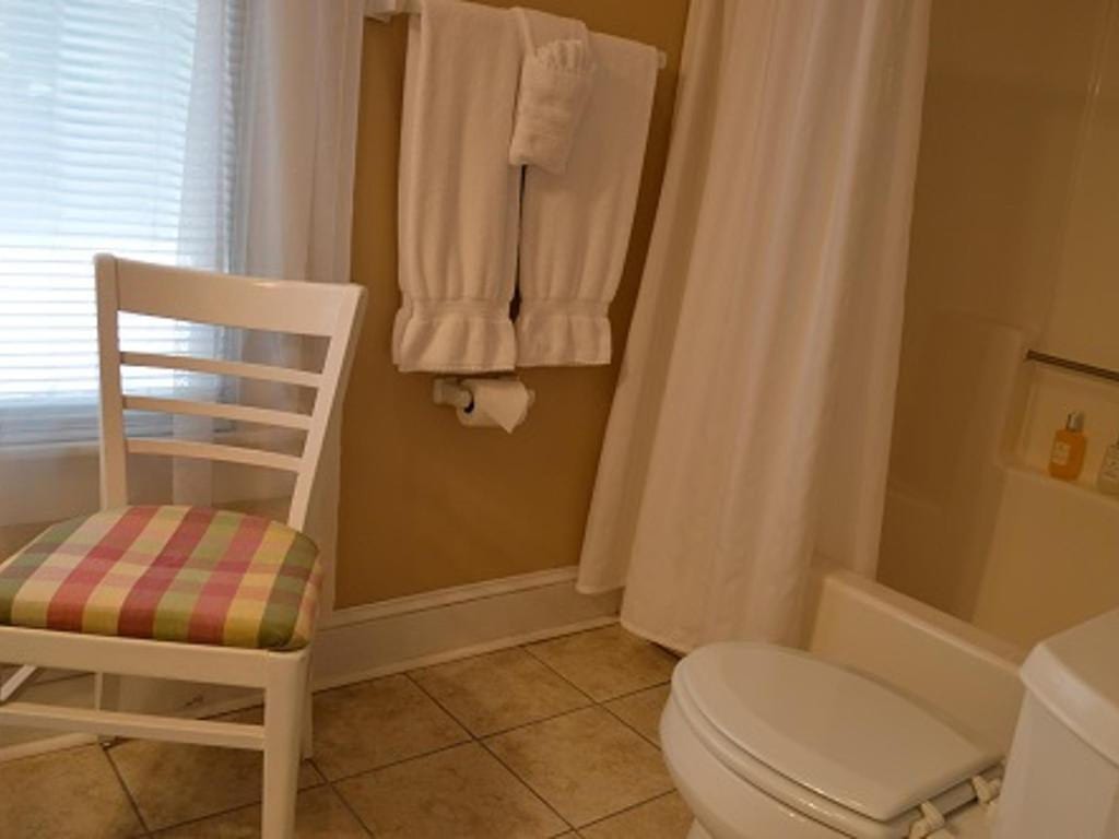 A white chair with a colorful checkered seat in a bathroom with a toilet, shower curtain, and towels hanging on a rack.
