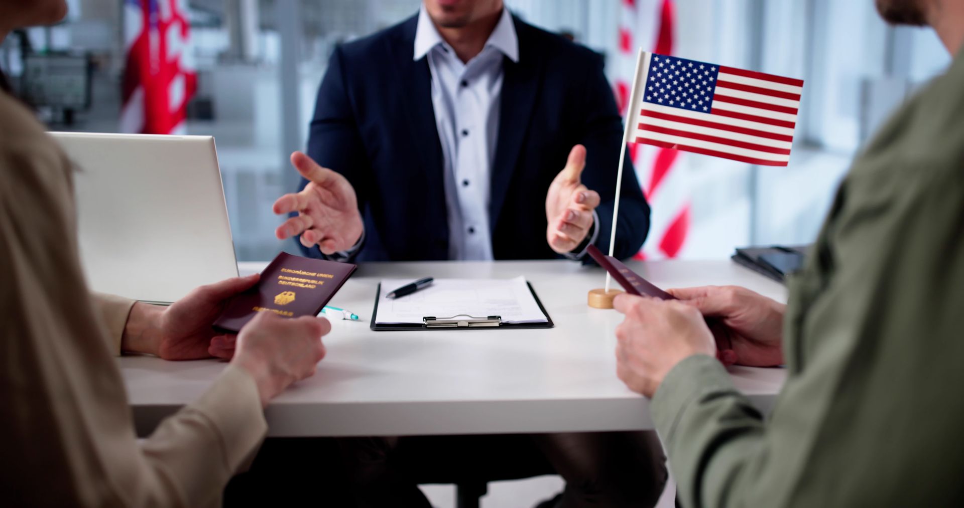 Two people discuss a passport at a desk while a small U.S. flag stands nearby.
