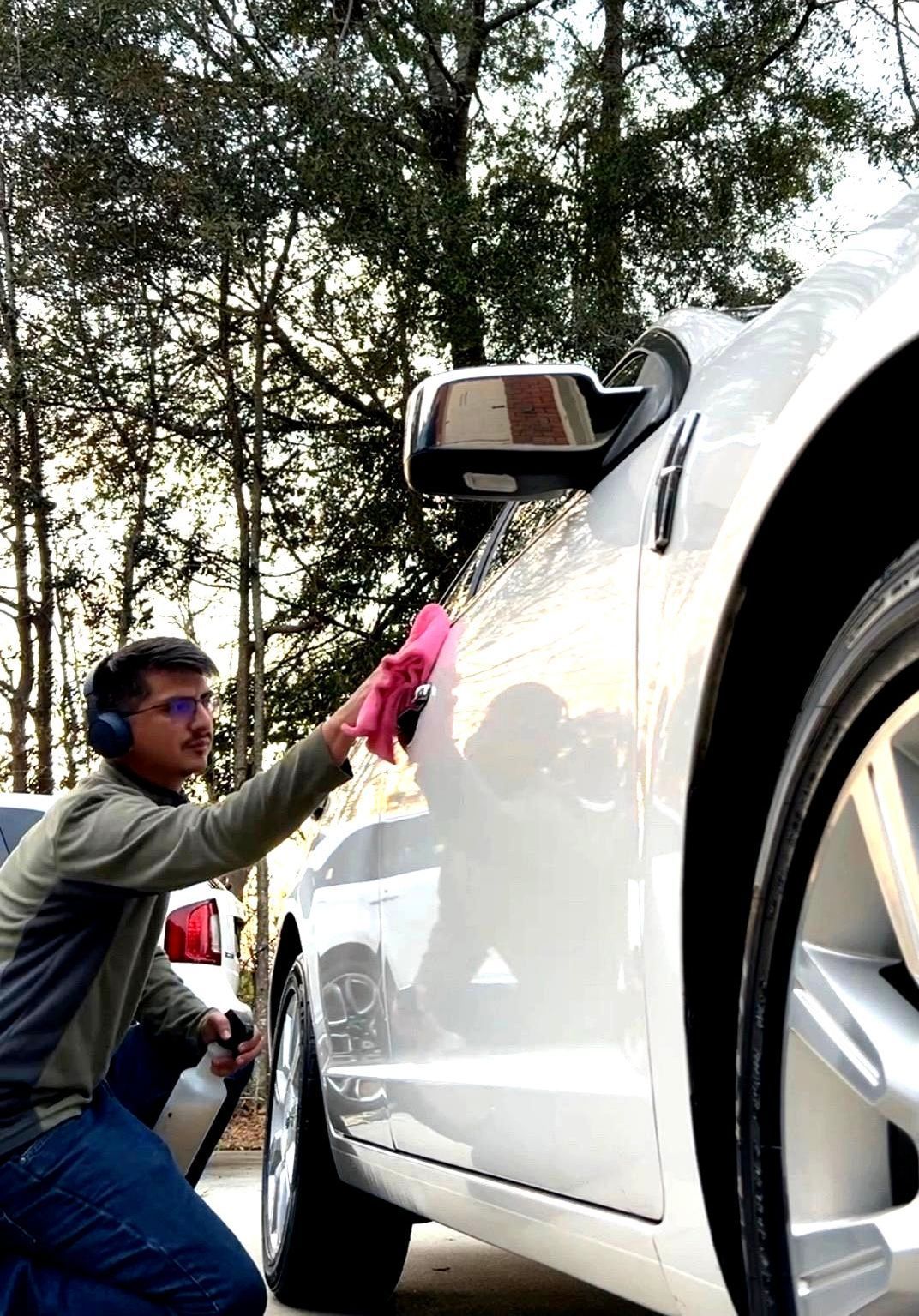 Person wiping down a silver car with a pink cloth outside; wearing safety glasses and ear protection.