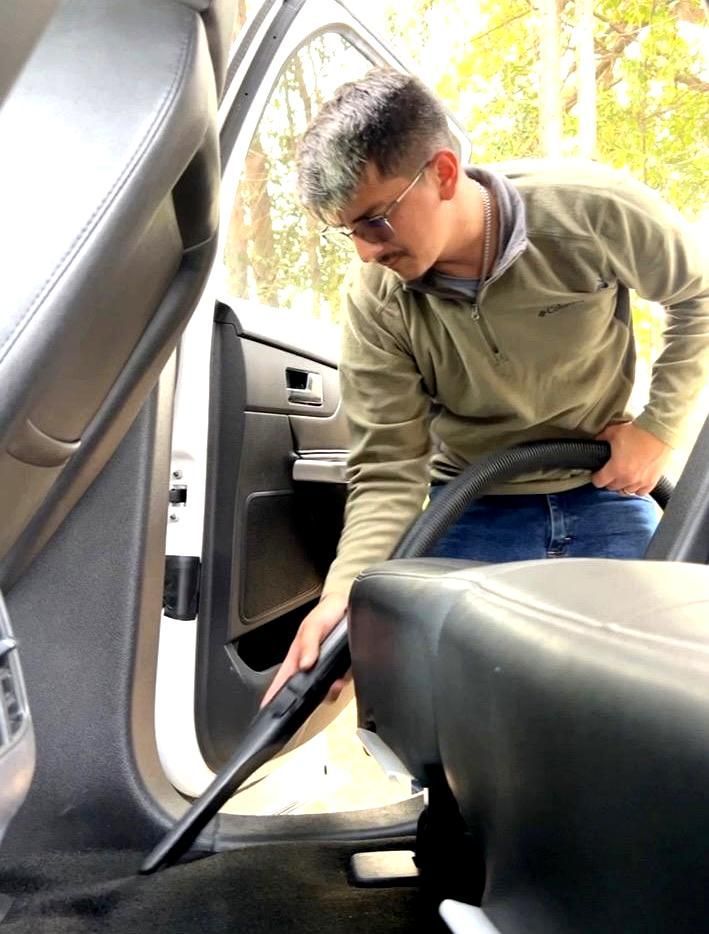Man vacuuming the interior of a car. He wears glasses and a green jacket, outdoors.
