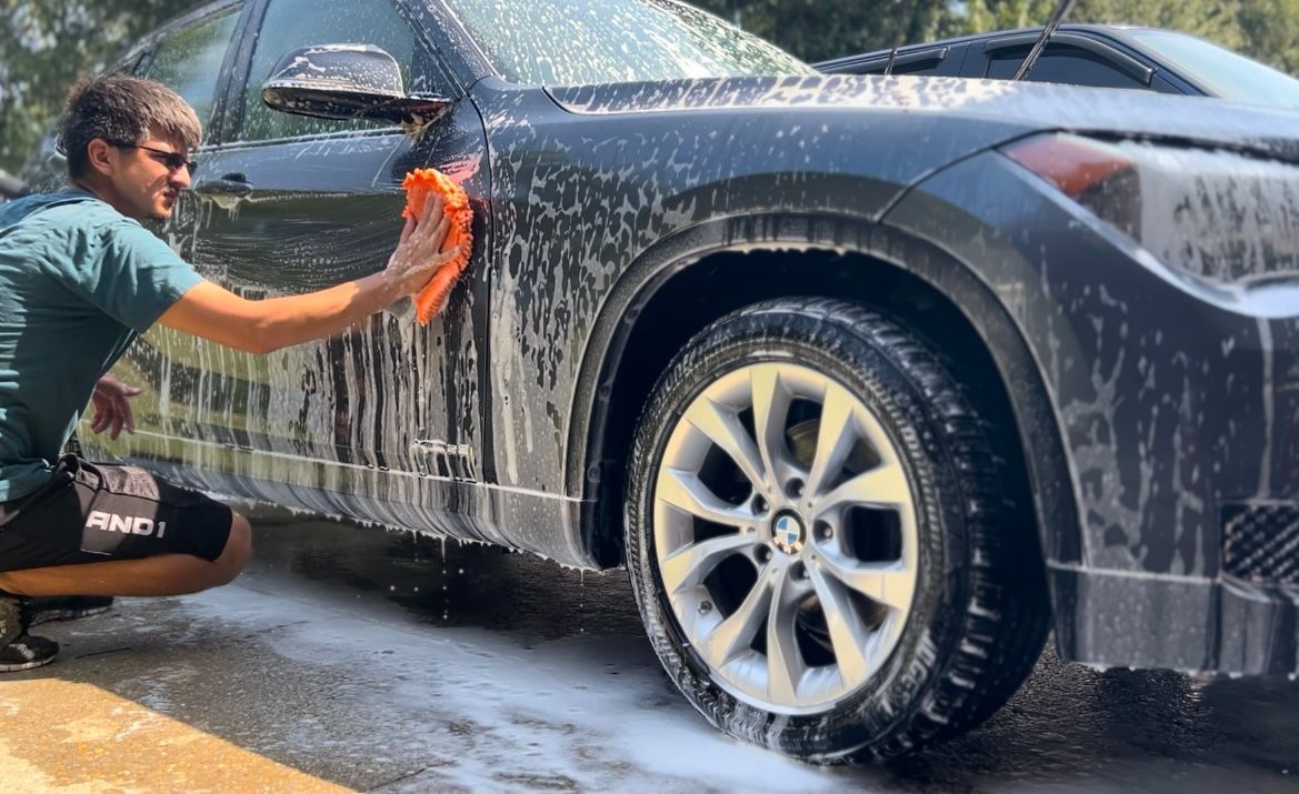 Person washing a dark-colored car with a soapy sponge outdoors on a sunny day.