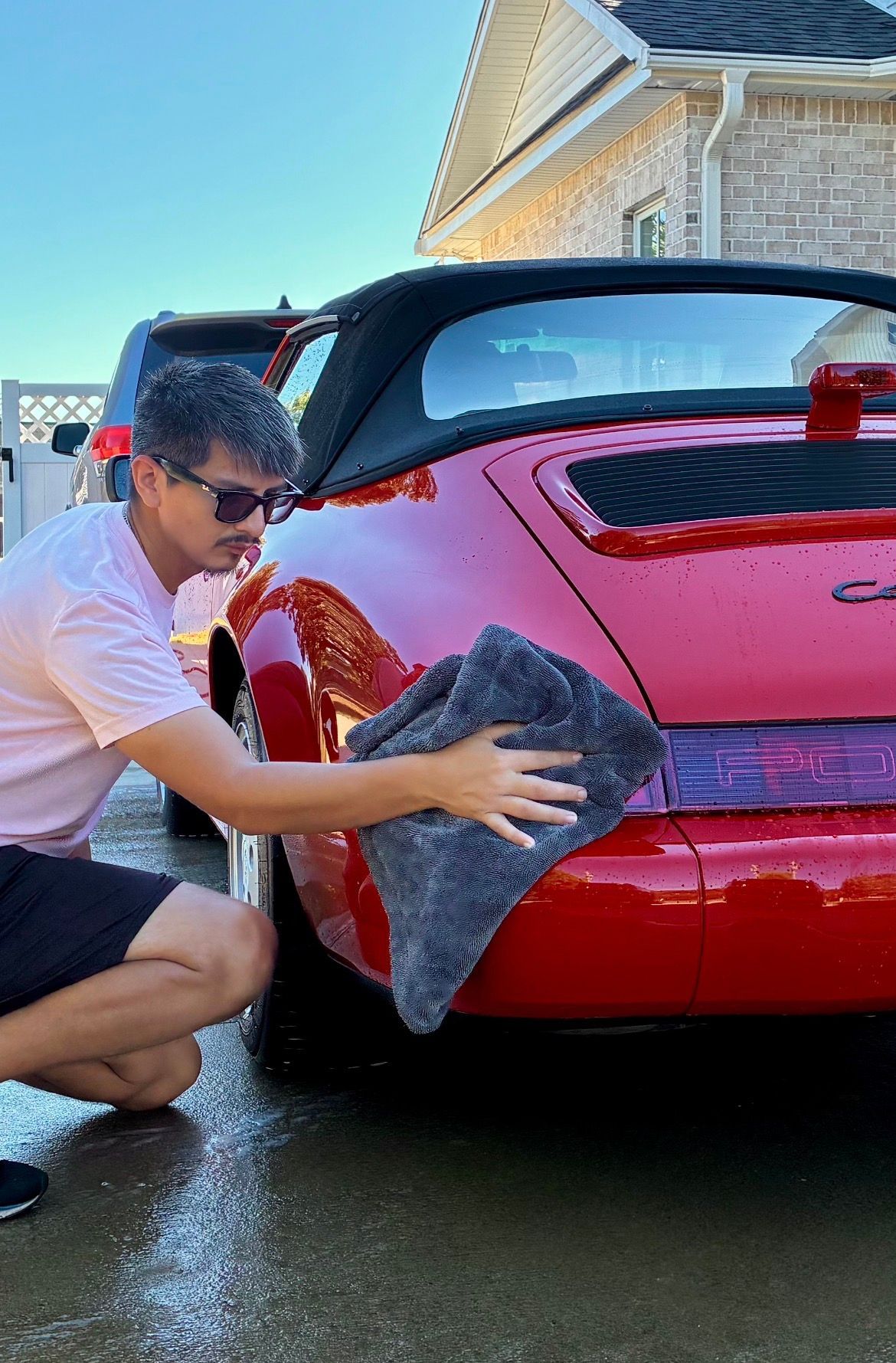 Man drying a red convertible car with a gray cloth. Outdoors, sunny.