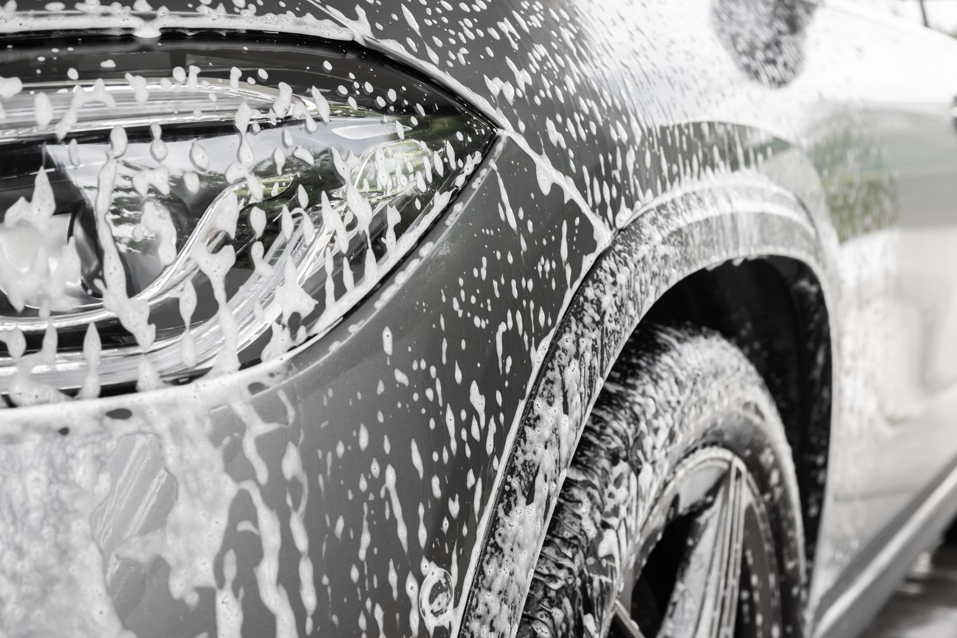A car being washed with foamy soap, close-up view of the side, headlight, and tire.