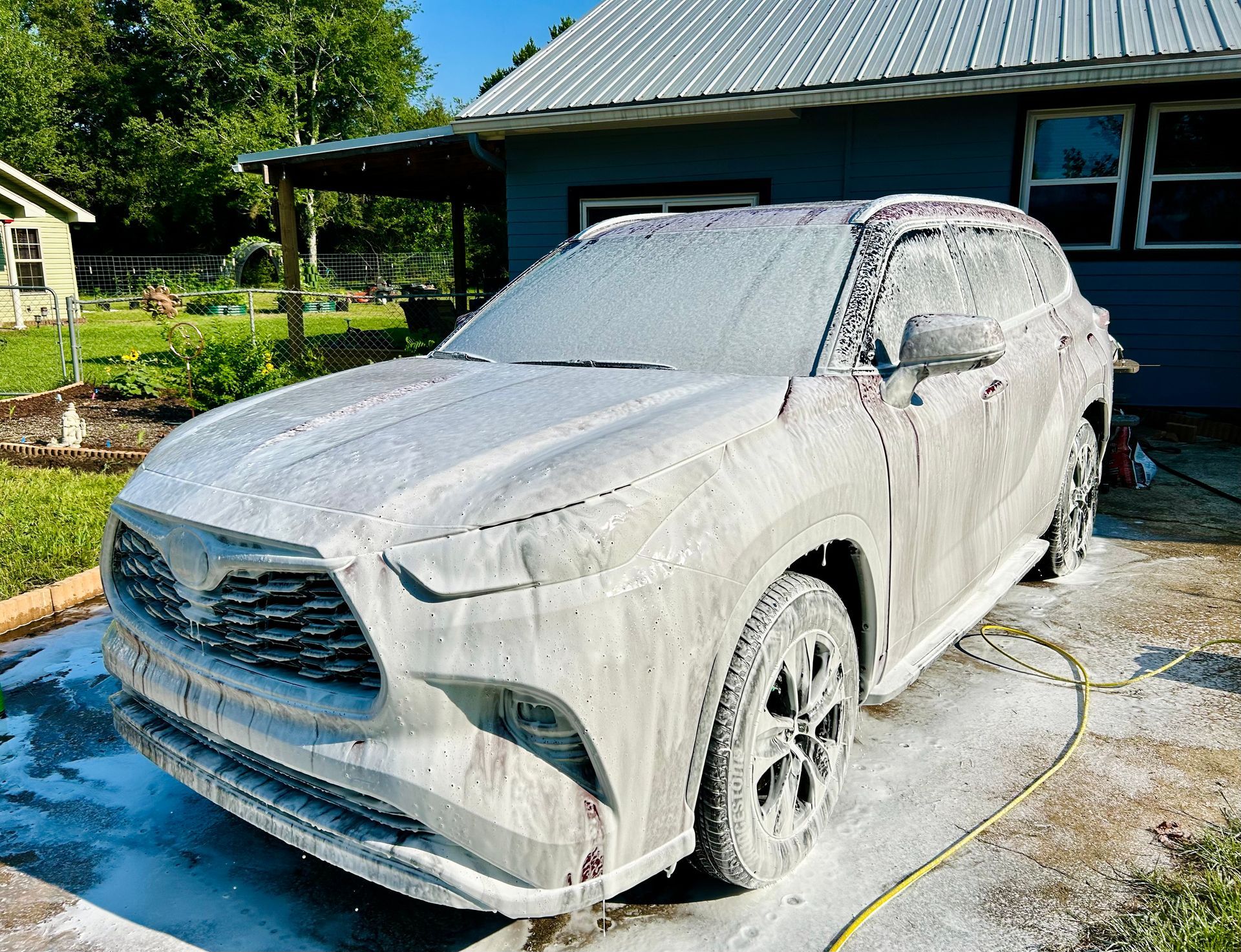 White SUV covered in foamy soap suds, parked in front of a blue house and yard, during a car wash.