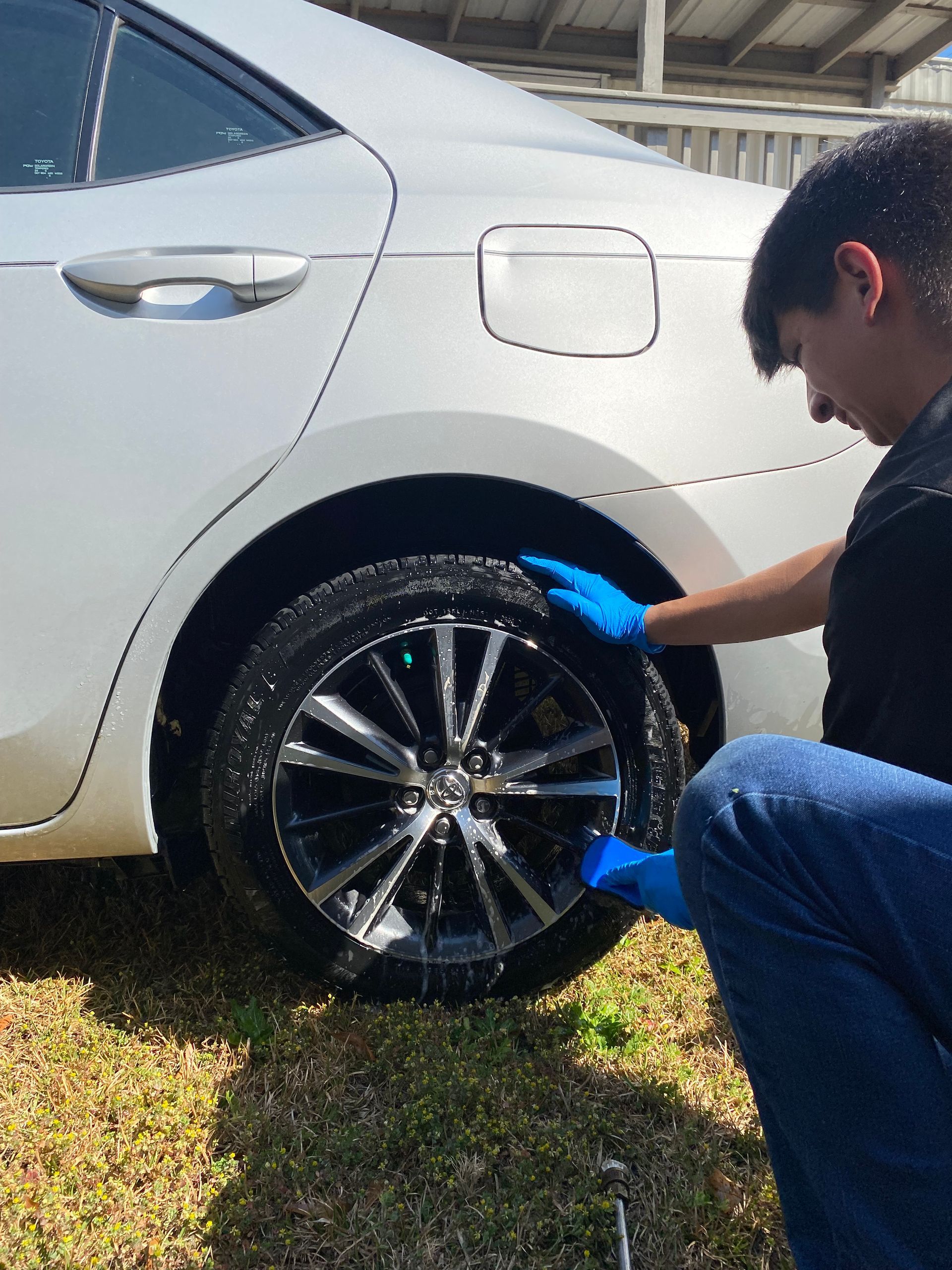 Person wearing blue gloves cleans a white car's tire with suds outdoors.