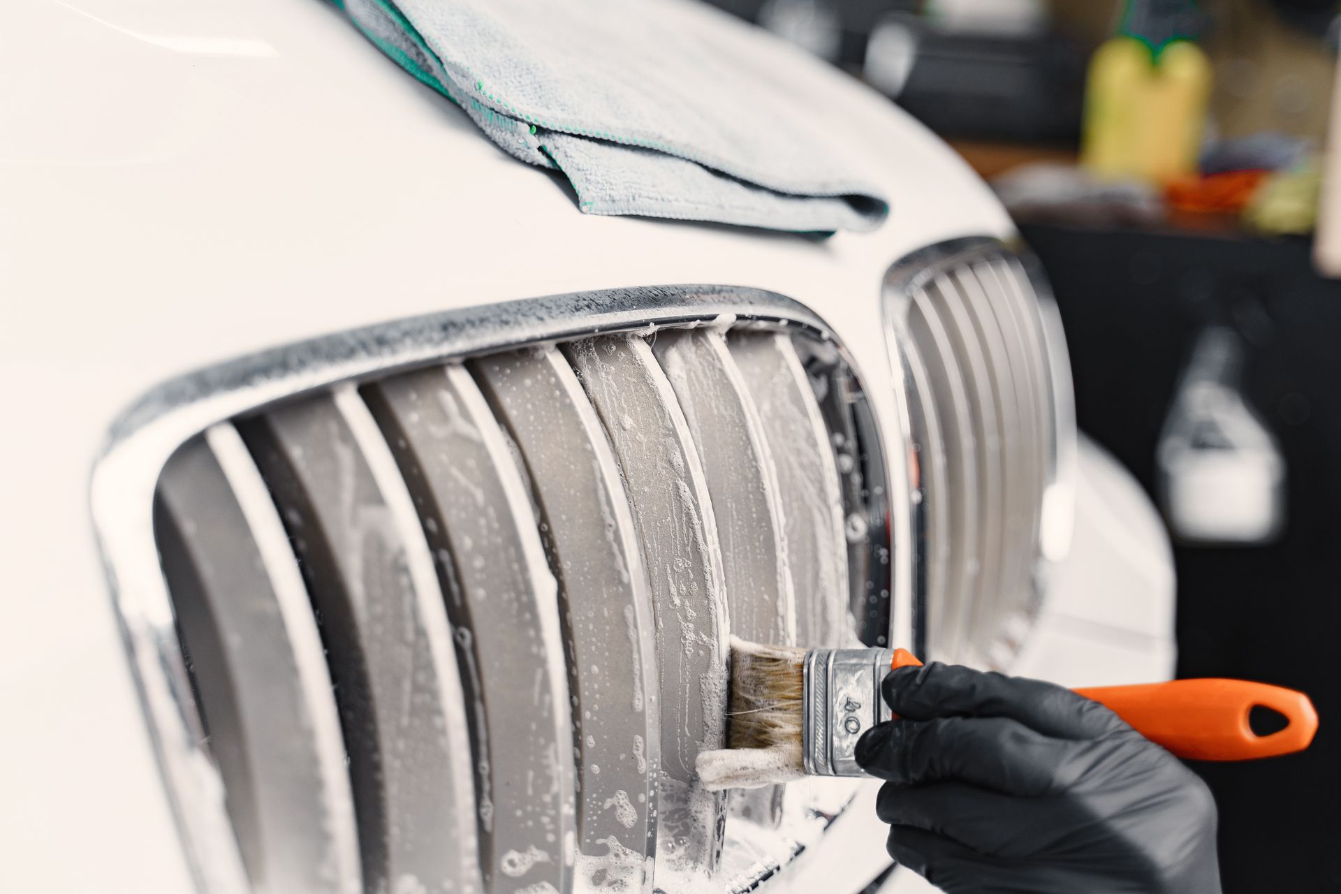 Person cleaning a car's white grill with a brush and soapy solution, using a black glove.