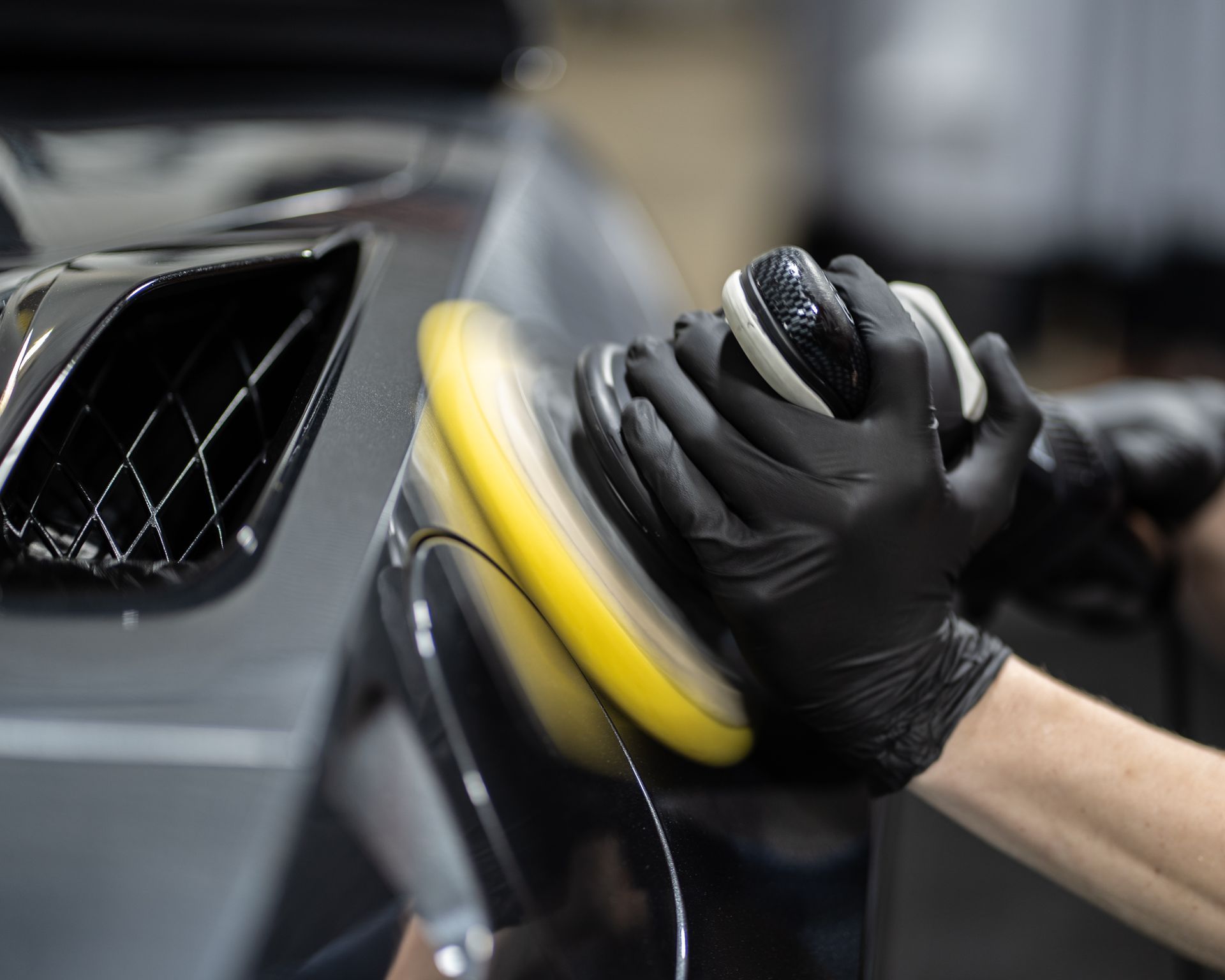 Person wearing black gloves polishing a dark-colored car panel with a yellow buffing pad.