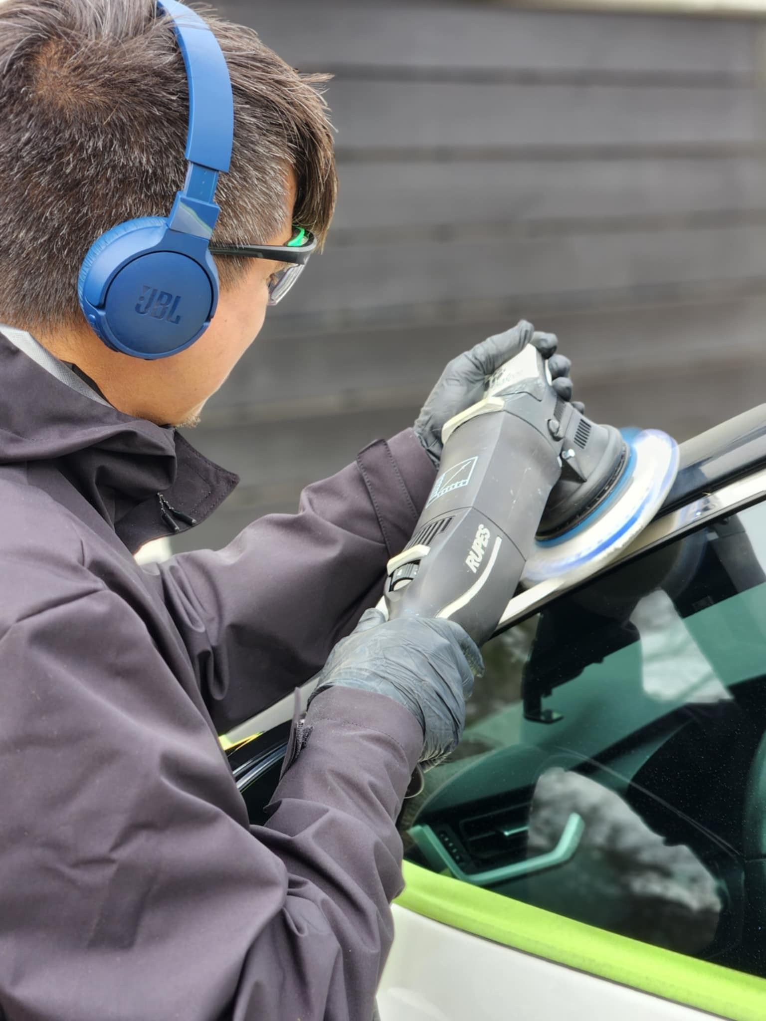 Person using a polisher on a car's window, wearing gloves, headphones, and safety glasses.