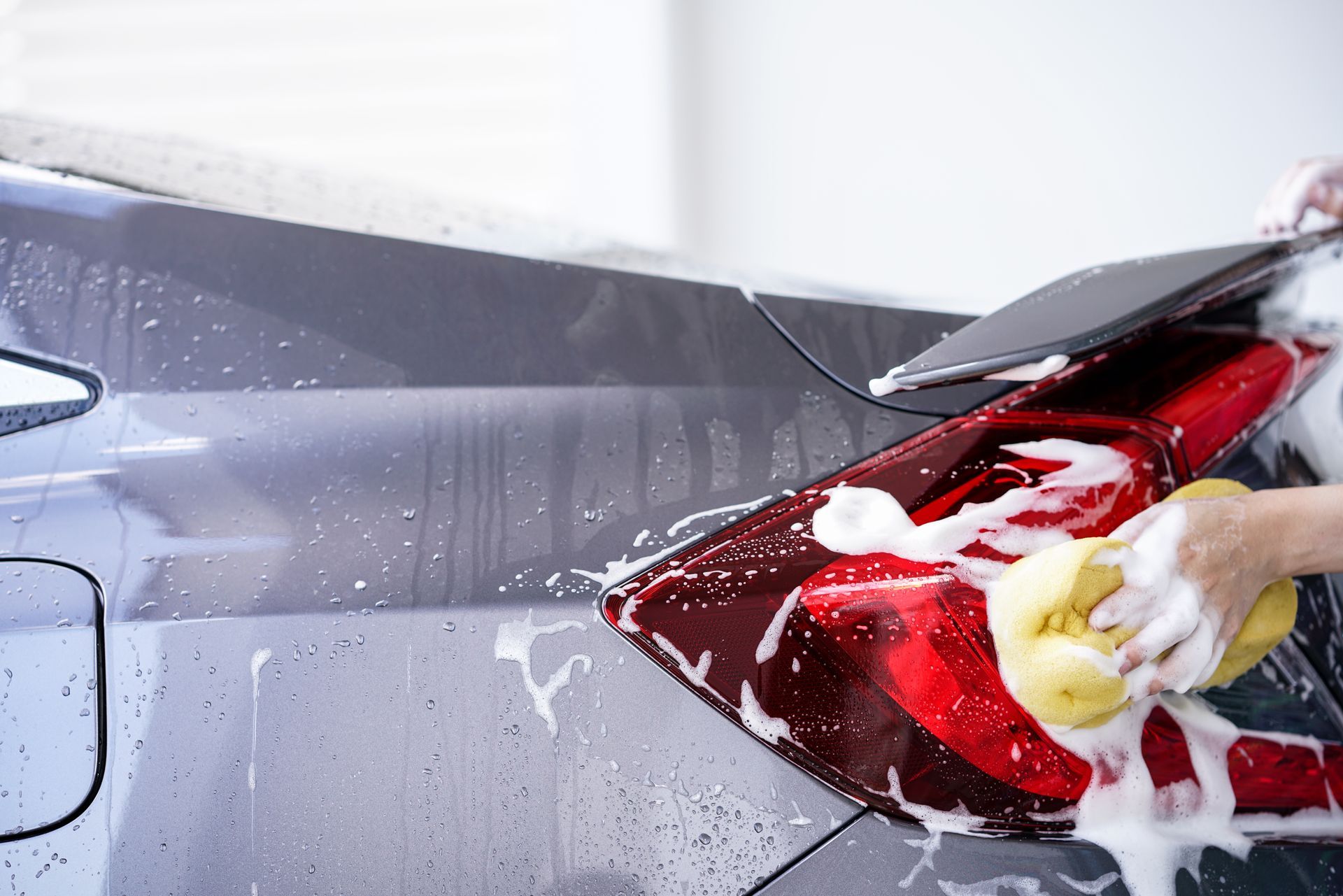 Washing the tail light of a gray car with a soapy sponge.