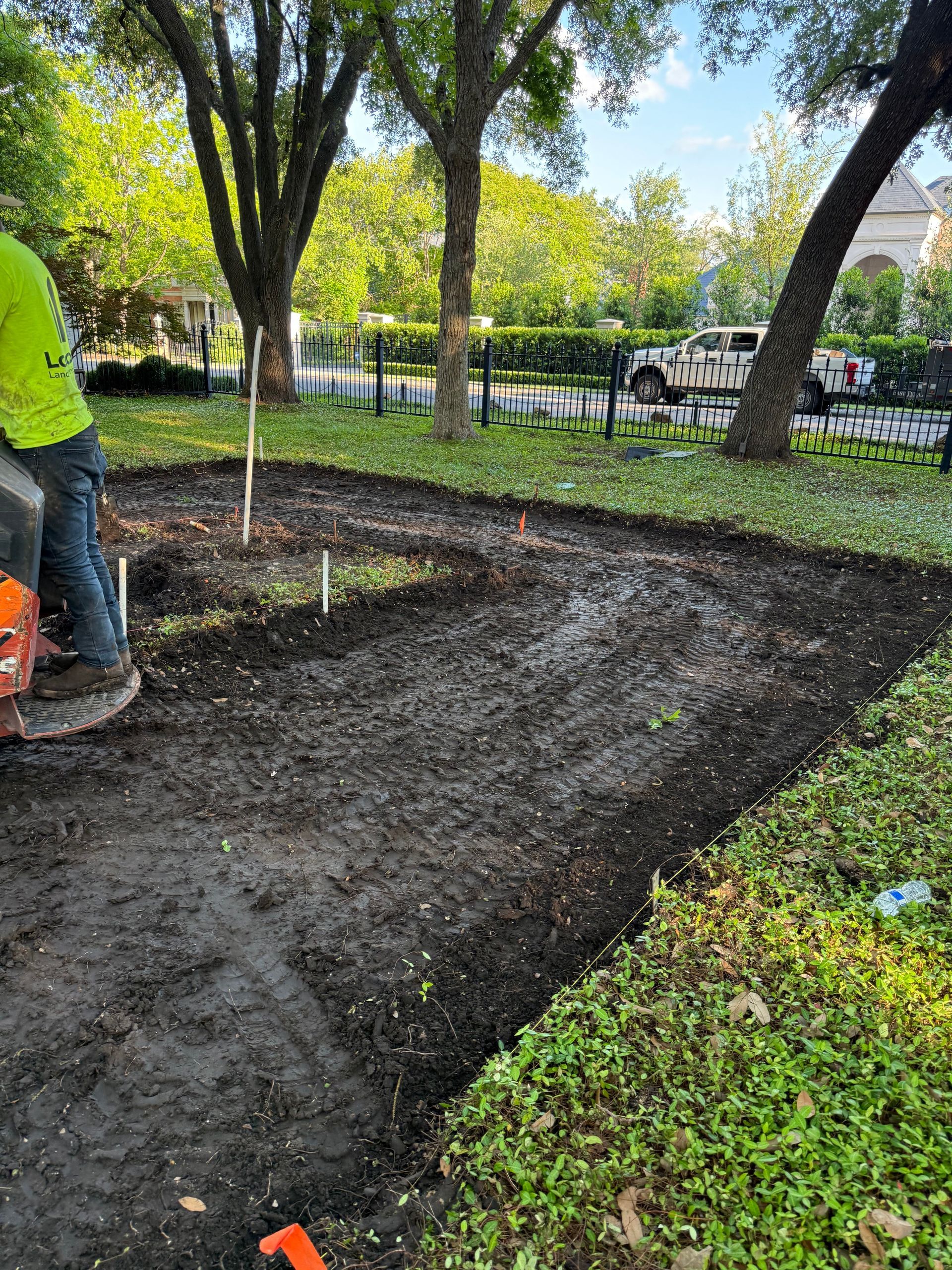 Man operating machinery in a newly tilled garden bed, surrounded by green foliage and trees.