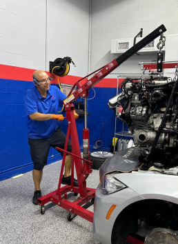 Mechanic using a red engine hoist to lift an engine from a silver car in a shop with blue and red walls - Bimmerfix